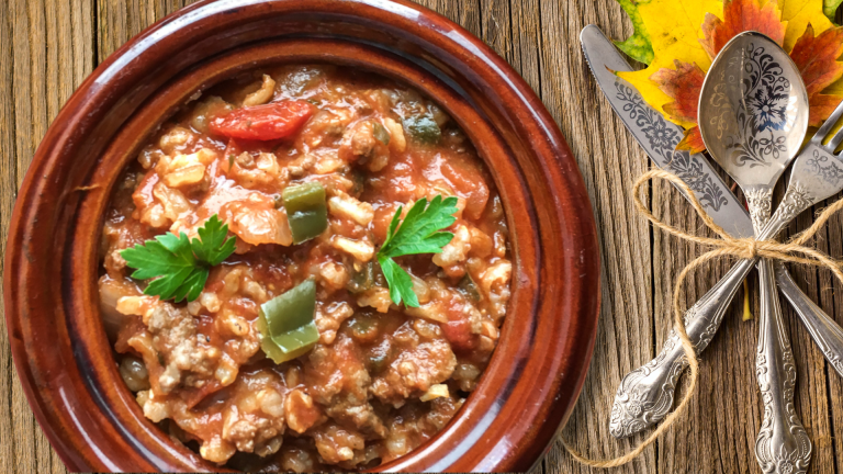 A rustic bowl filled with Weight Watchers-friendly sausage and pepper soup, garnished with parsley, sitting on a wooden table. Beside the bowl is a set of ornate silverware tied with twine, accompanied by colorful autumn leaves. Perfect for a cozy, healthy meal.