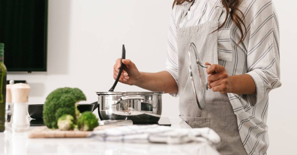 A young woman, wearing an apron over a striped shirt, cooks a healthy dinner with cookware that is free of forever chemicals.