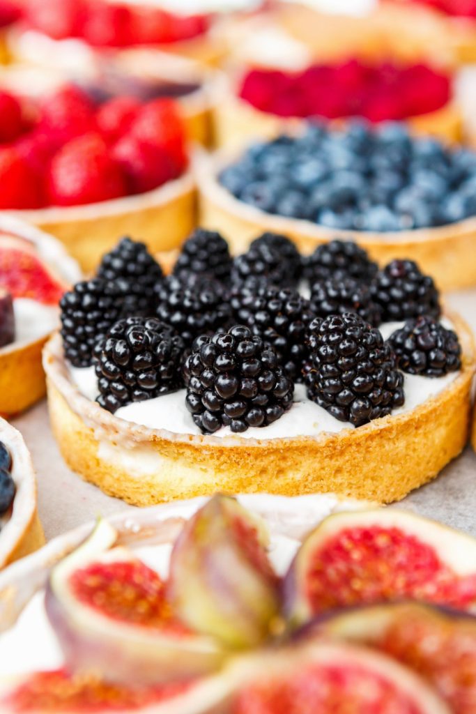 A close-up view of a dessert tart with blackberries on top among several other dessert tarts with fresh fruit.