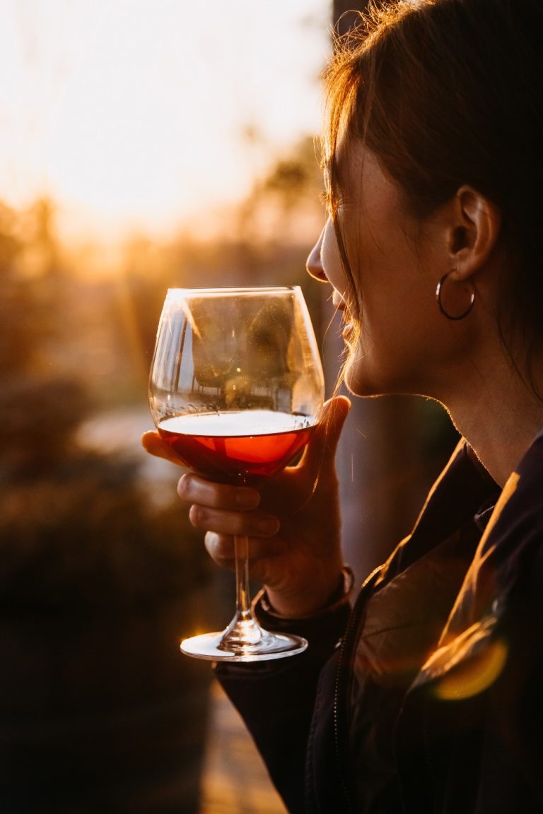 A woman holding a glass of reddish wine and leaning in her window as she stares out at the gentle sunset.