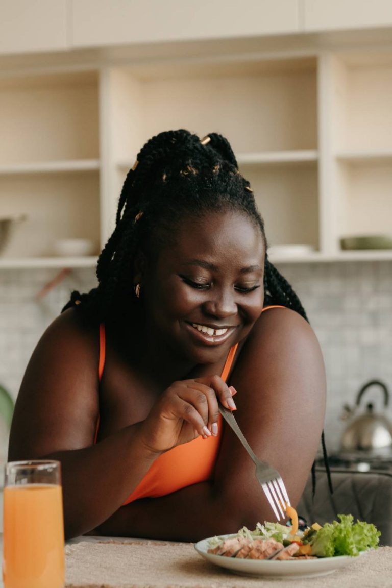 A woman wearing an orange top enjoying a healthy lunch and juice at home. She's laughing with her fork in her hand.