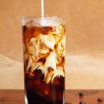 A glass full of cold brew coffee on ice. Someone is pouring cream into the glass and there are stray beans on the table.