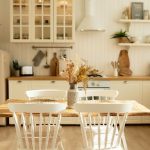 A cozy, organized kitchen with lots of warm, natural light, a small wooden dining table, and a cream colored fridge.
