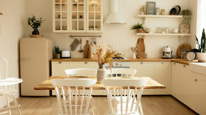 A cozy, organized kitchen with lots of warm, natural light, a small wooden dining table, and a cream colored fridge.